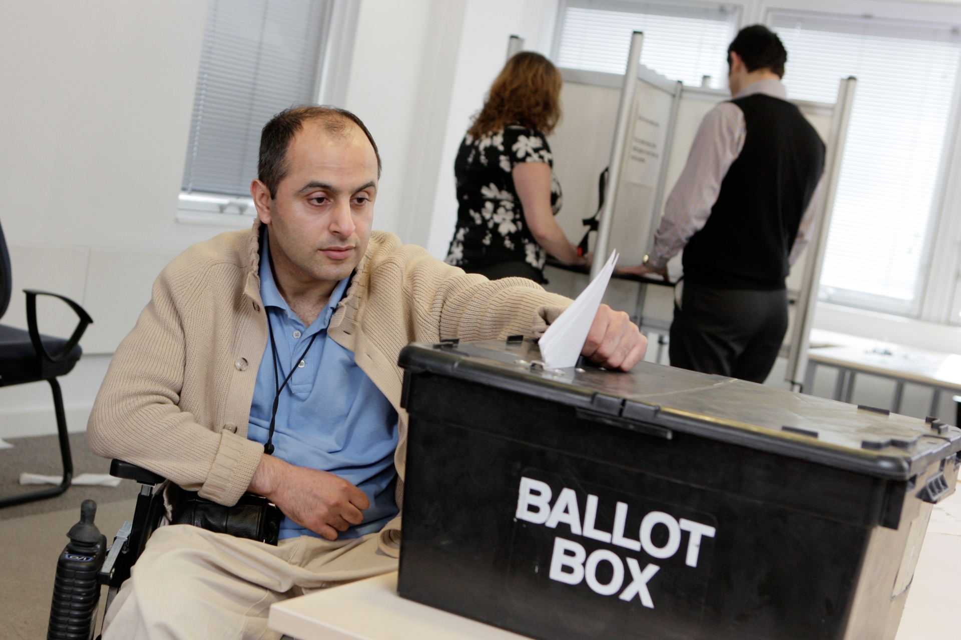 A man casts his vote in a ballot box. Behind him, two people mark their ballots.